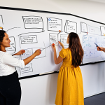 **

A team of diverse professionals (developers, marketers, designers) collaboratively sketching on a large whiteboard filled with wireframes and user flows. The setting is a bright, modern office space in Amsterdam. Focus on the energy and interaction, highlighting sticky notes with Dutch phrases like "Kernfunctionaliteit" and "Feedback".

**
